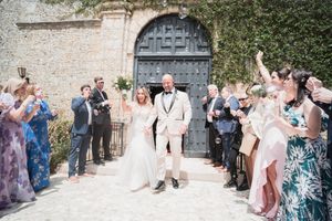 Bride and groom exit church ceremony through confetti shower, guests celebrating outdoors at sunny European wedding venue.