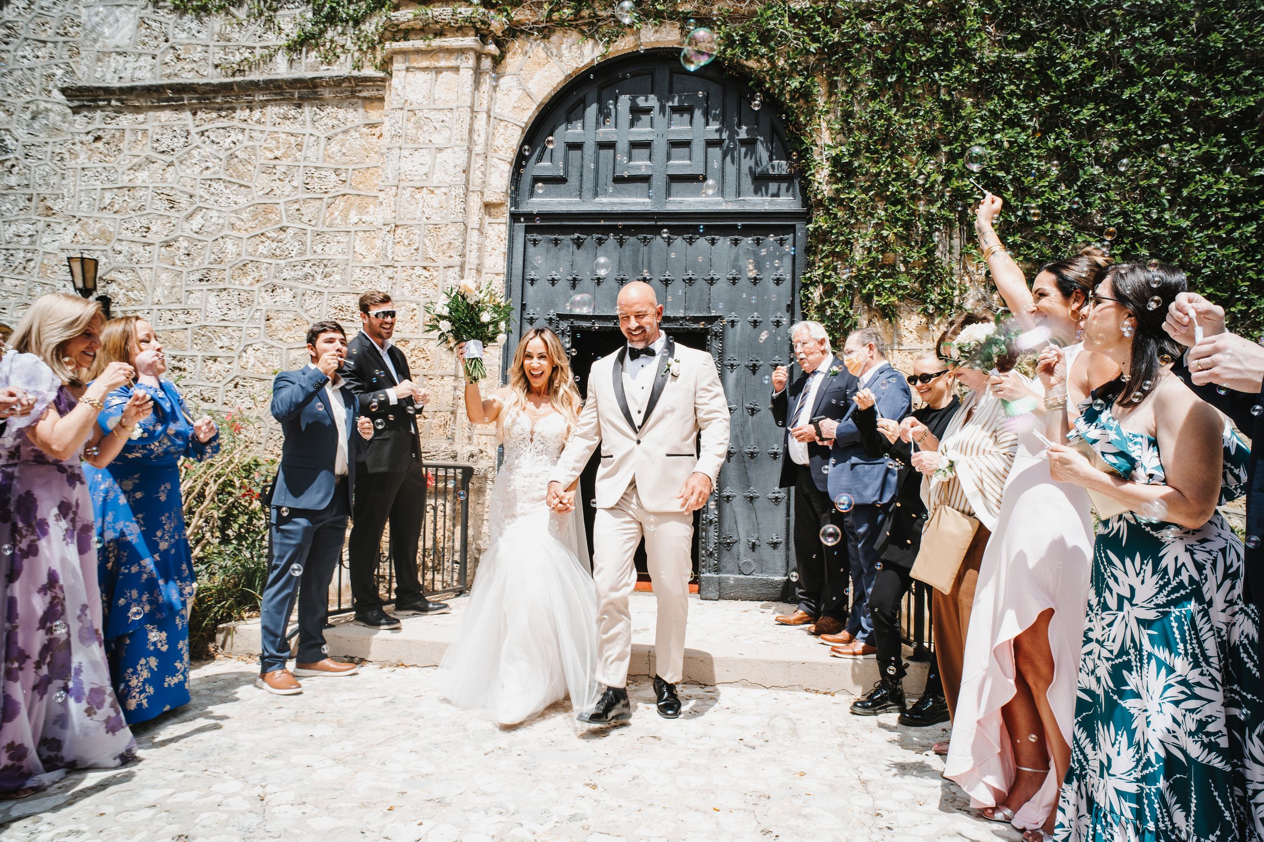 Wedding guests throw confetti at newlyweds outside a historic stone church with ivy-covered walls.