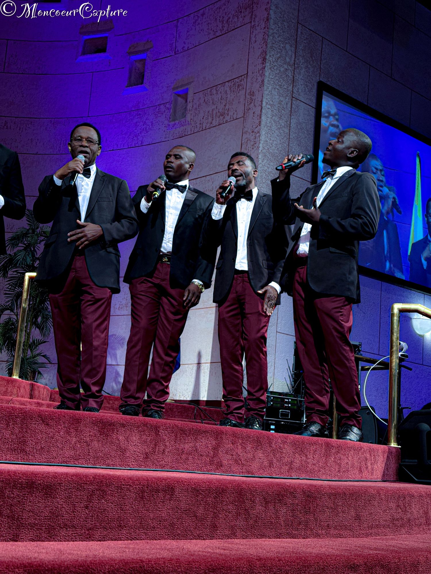 Male quartet in matching burgundy pants and black tuxedos performs on stage with microphones.