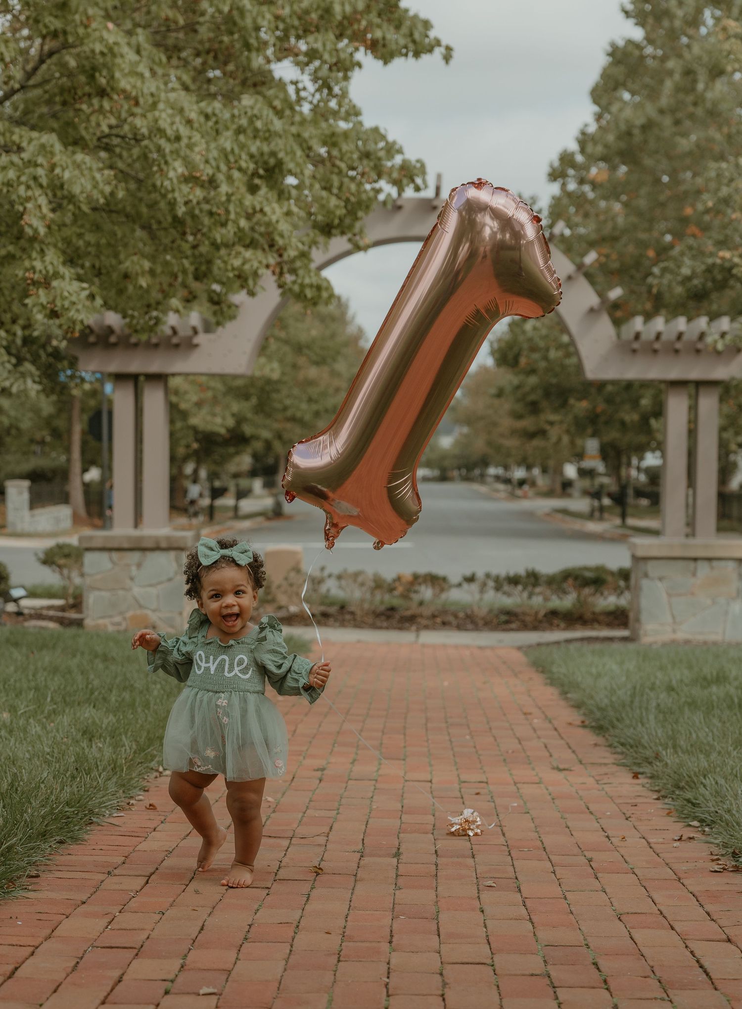 A joyful toddler in a mint green dress plays with a large number one balloon on a brick walkway.