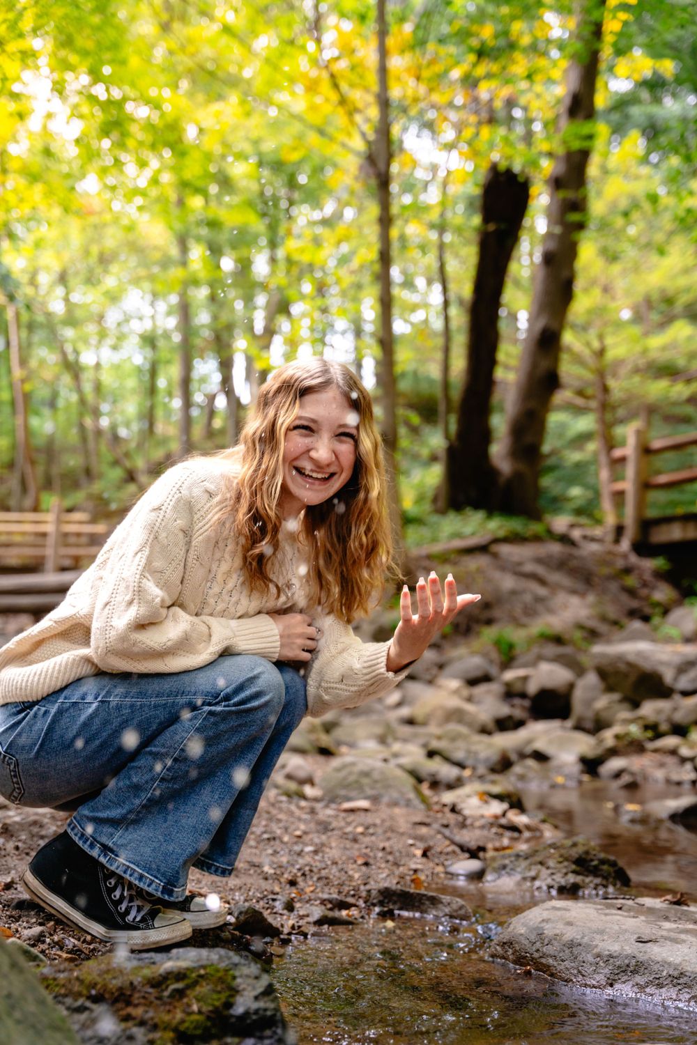 Person in cream sweater crouching by stream in autumn woods with joyful expression. Senior photography