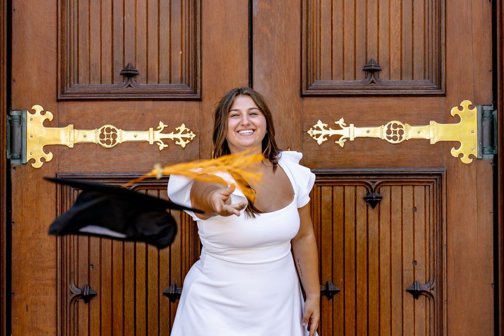 Graduate in white dress stands playfully between two wooden church doors adorned with vintage iron hardware and lighting.