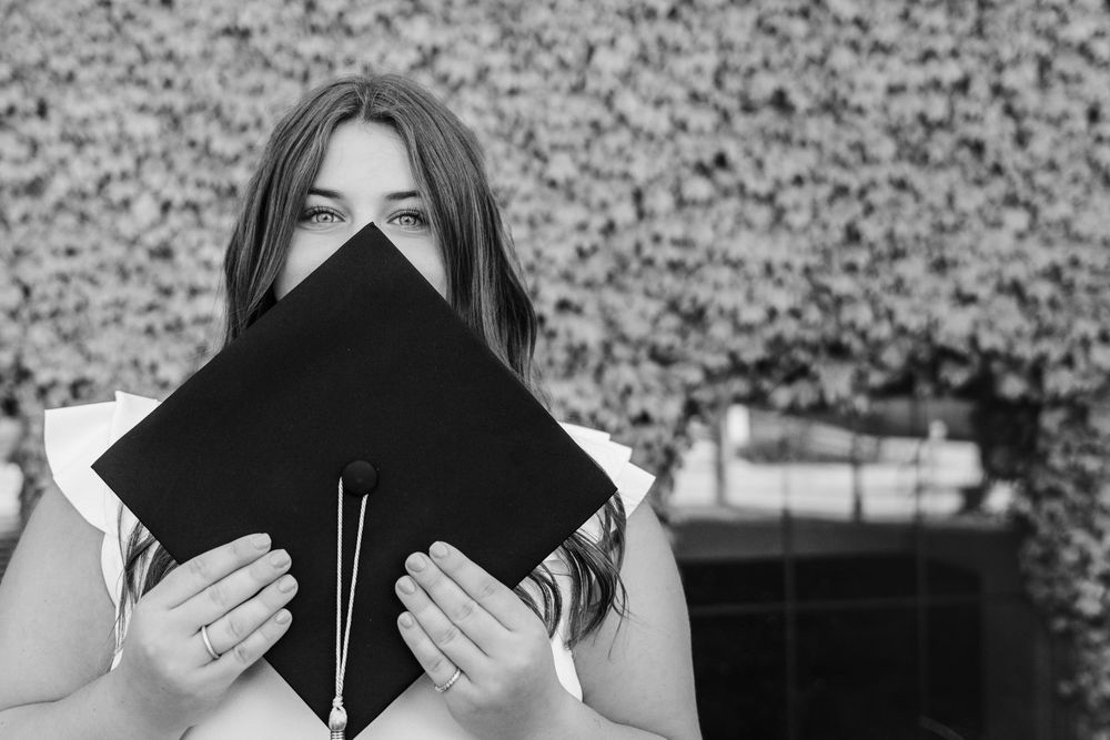 Graduate in cap and gown holds their graduation cap in front of their face in black and white portrait. Graduation from nursing school