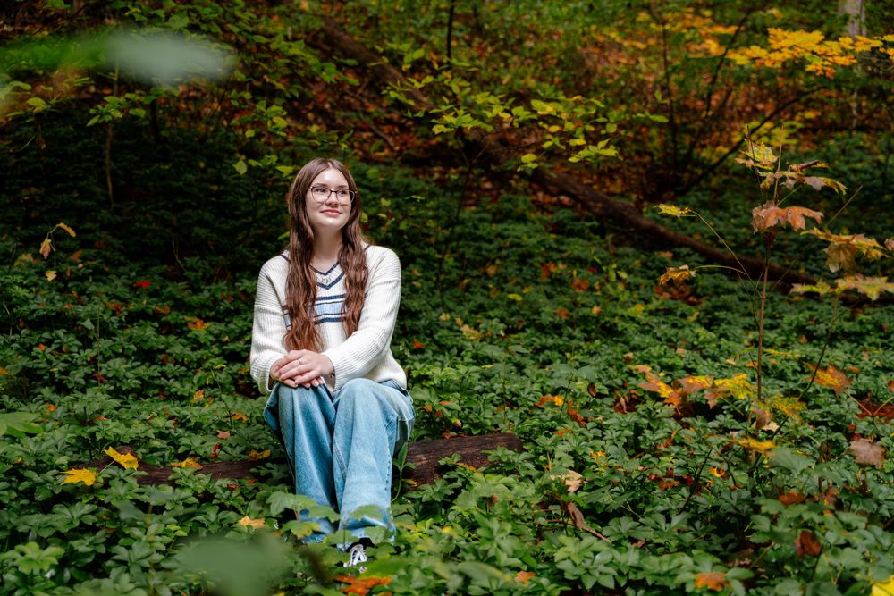 Someone sits on a mossy log surrounded by lush green foliage and autumn colors in a peaceful forest setting.