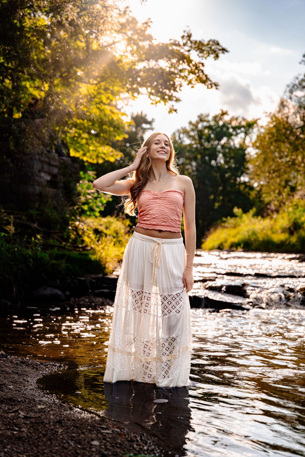 Young woman in pink top and white lace maxi skirt standing in a sunlit river surrounded by lush green trees.