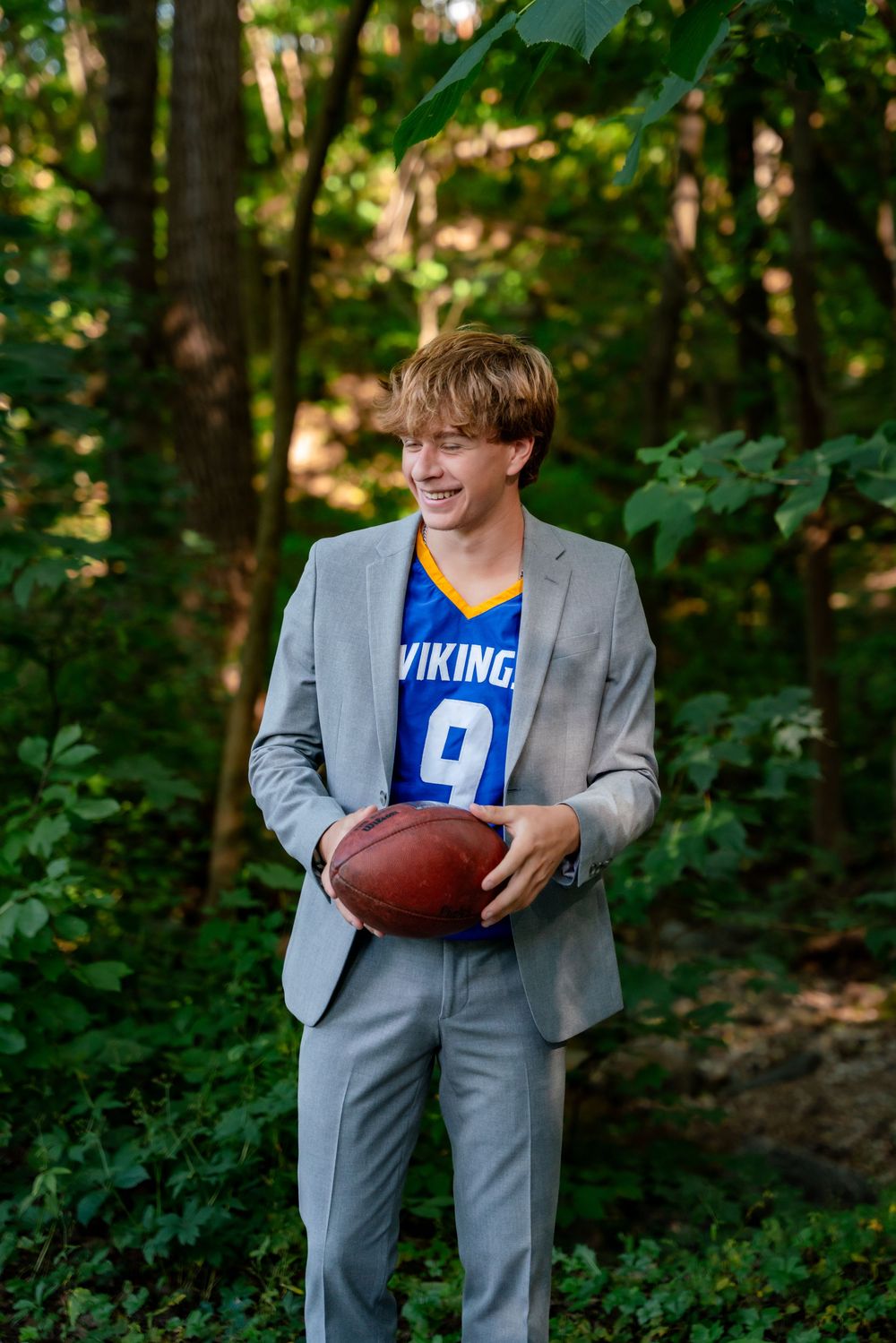Smiling teen in gray suit and Vikings jersey holding football outdoors in lush green forest setting.