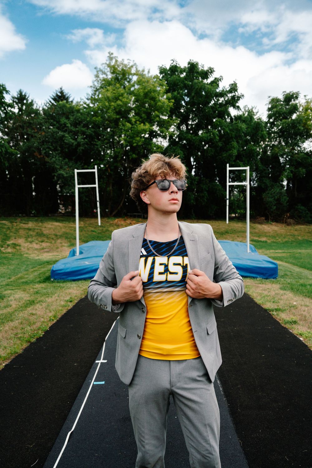 Young man in gray suit jacket over yellow West jersey and sunglasses poses confidently on a school track field.