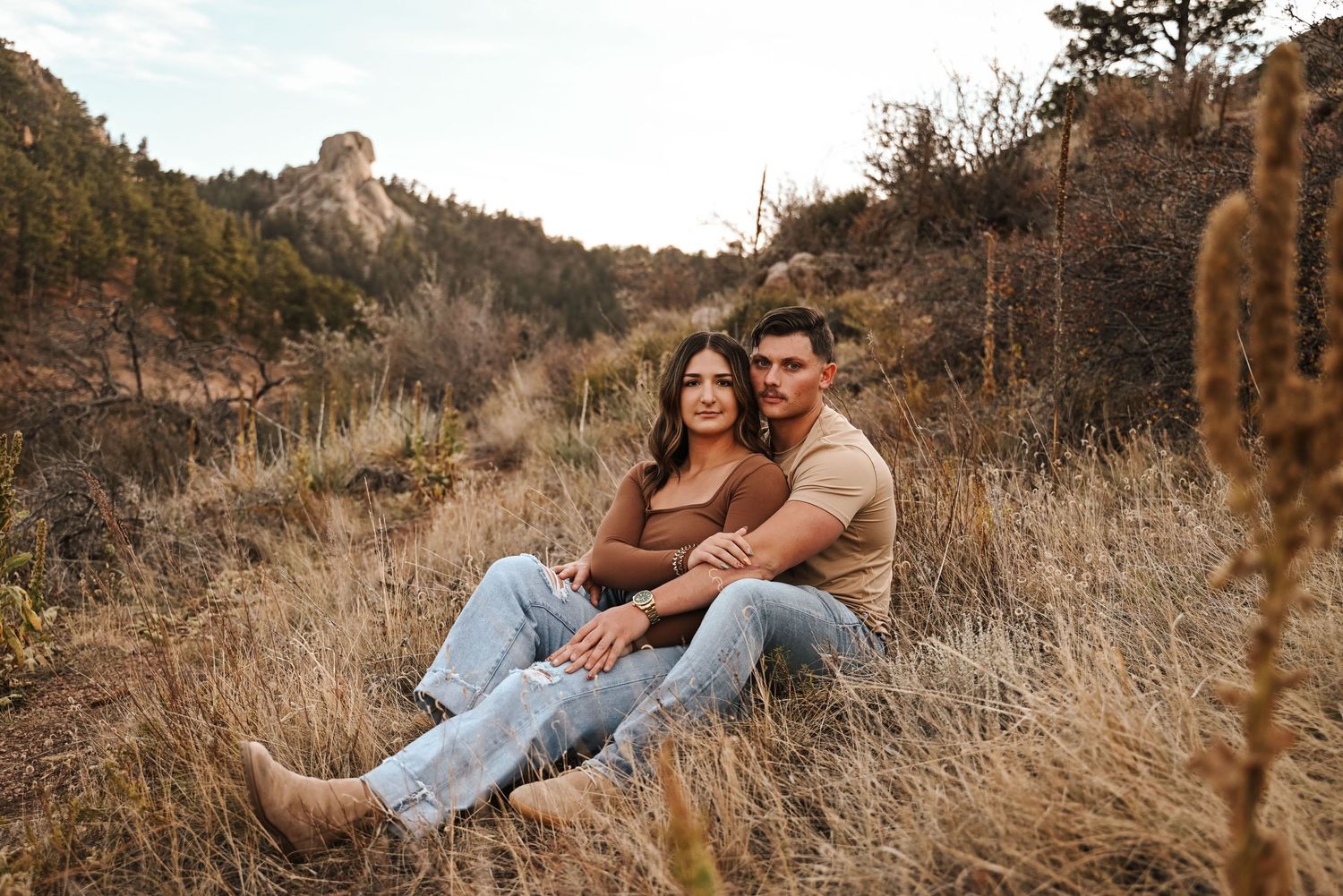 Couple sitting together in tall grass with mountain scenery.