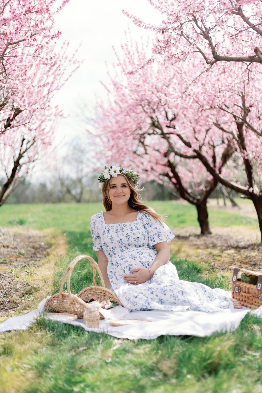 Springtime picnic setup under blooming cherry blossom trees with white blanket, woven baskets and delicate floral details.