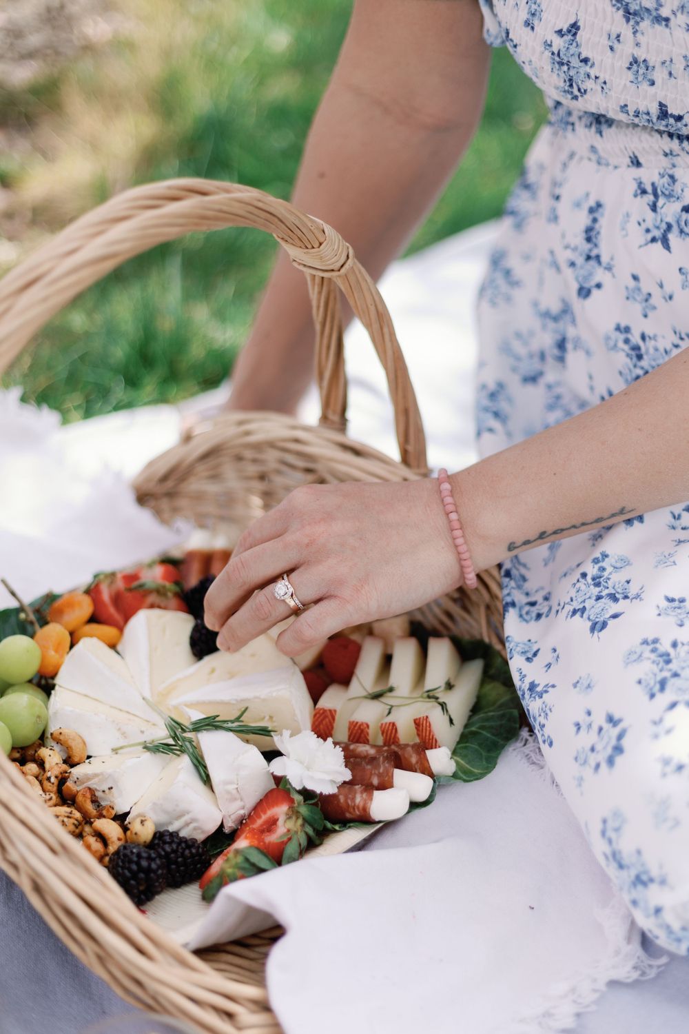 Someone wearing a floral dress reaches into a wicker picnic basket filled with cheese, fruits and charcuterie on a blanket.