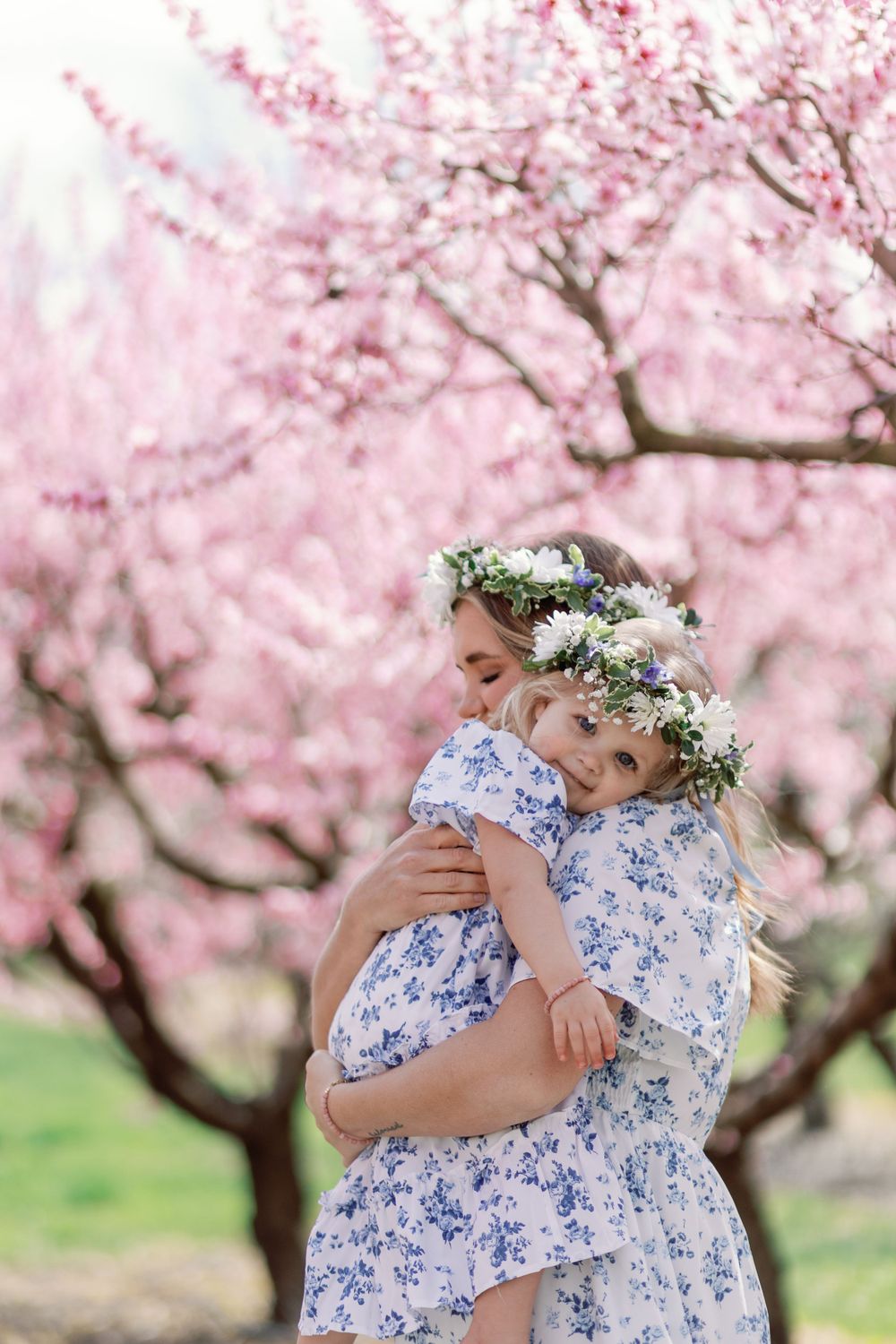 Two people in matching blue and white floral dresses share a tender embrace under blooming pink cherry blossom trees in spring.