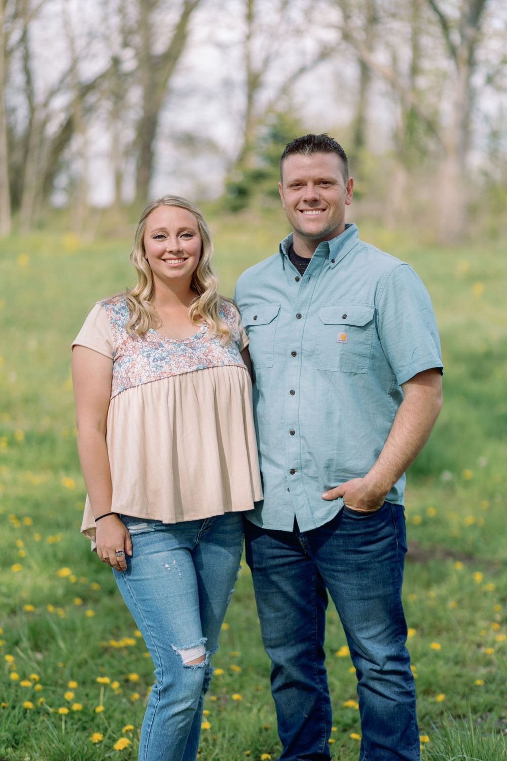 A couple stands together outdoors in a spring meadow with yellow wildflowers and trees in the background.