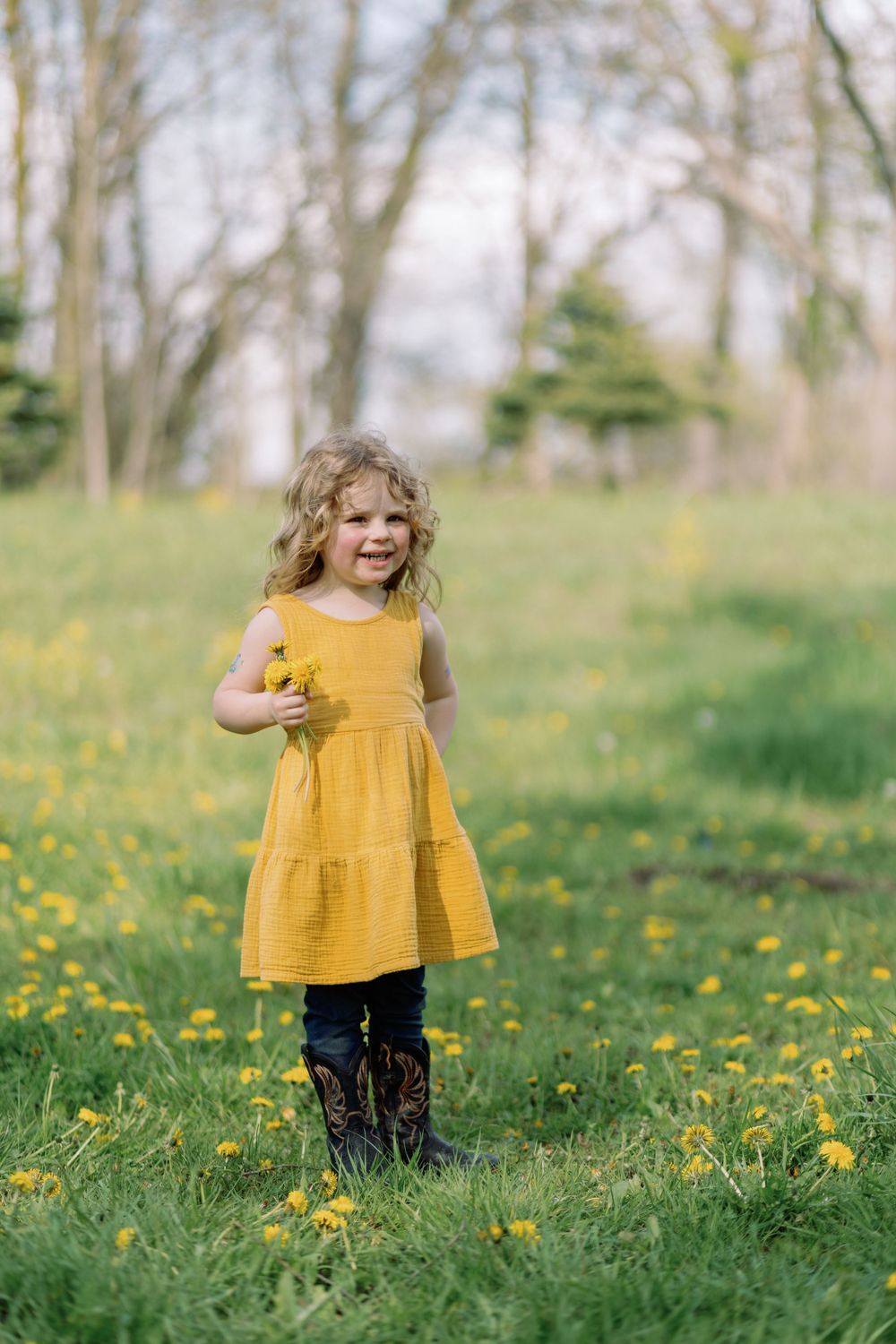 A child in a mustard yellow dress stands in a spring meadow filled with blooming dandelions.