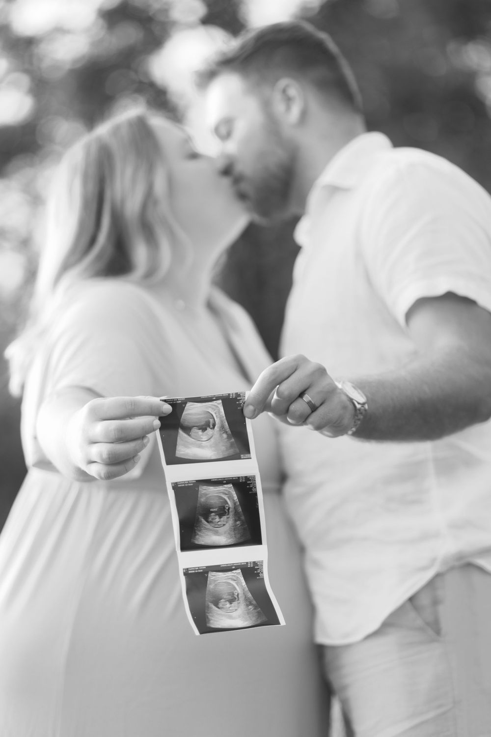 A couple shares an intimate moment while holding ultrasound photos of their expected baby in a black and white photo.