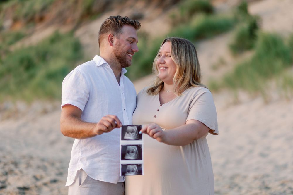 A couple on a beach shares a joyful moment while holding ultrasound photos in the late afternoon light.