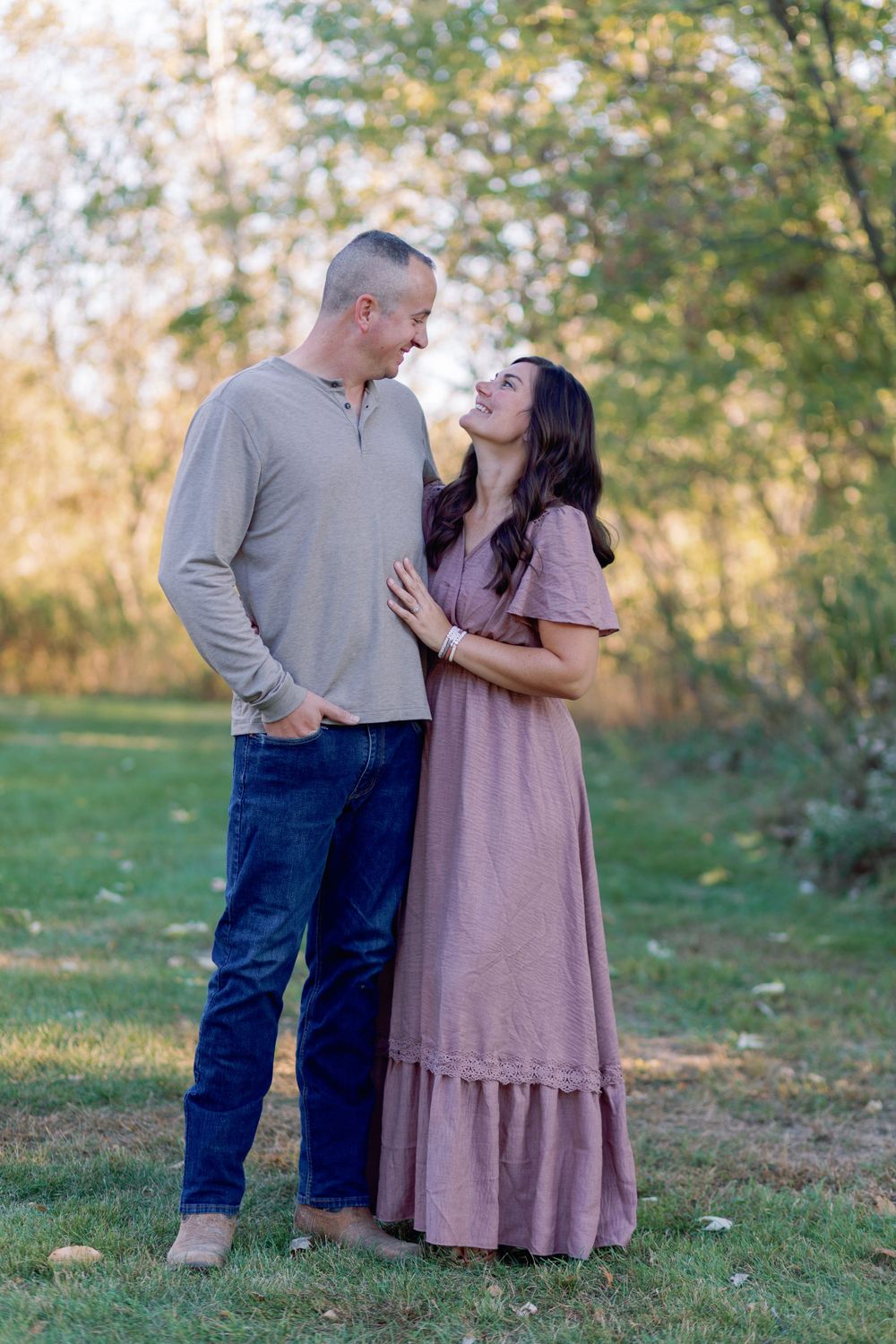 A couple shares a romantic moment outdoors, smiling at each other in a sunlit meadow with autumn trees in the background.