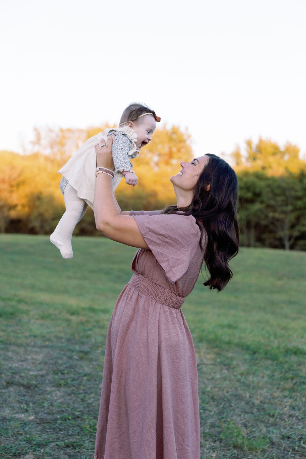 A parent and baby share a joyful moment outdoors on a grassy field with golden autumn trees in the background.