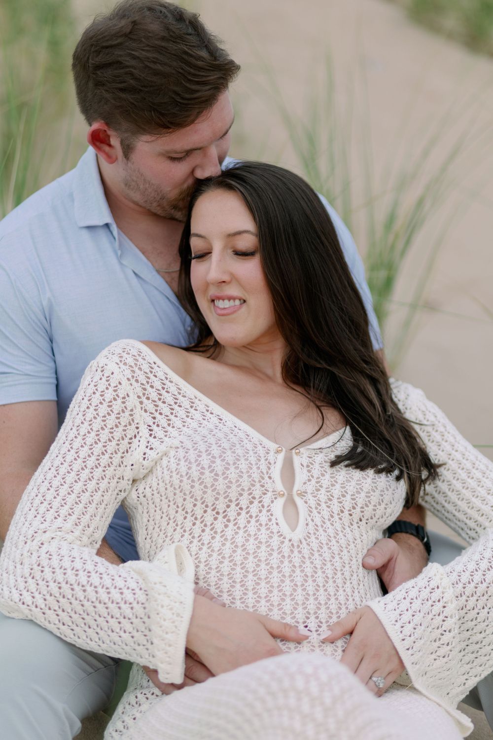 A tender romantic moment between a couple sitting together outdoors, with her wearing a white knit sweater, Oval beach, maternity shoot.