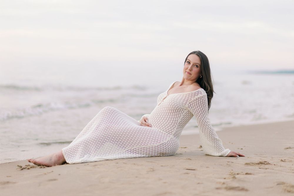 A pregnant woman in a white knit dress reclines on a sandy beach at sunset.
