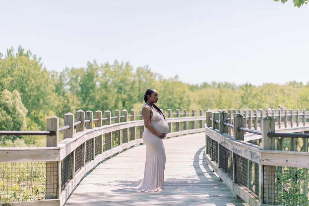 A person in a long white dress poses on a curved wooden boardwalk surrounded by lush summer greenery.