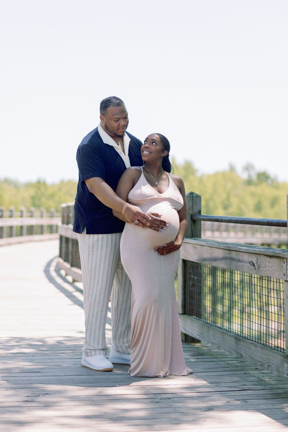 A loving couple embraces on a wooden boardwalk, with the expectant mother wearing a flowing white dress and her partner in navy.