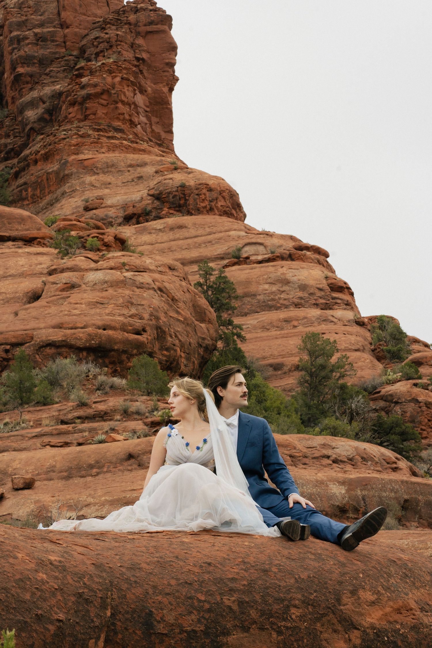Wedding couple sitting on red rock formations in Sedona with dramatic landscape backdrop.