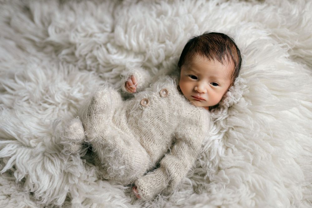 A newborn baby in a cozy knit onesie lies peacefully on a fluffy white fur rug.