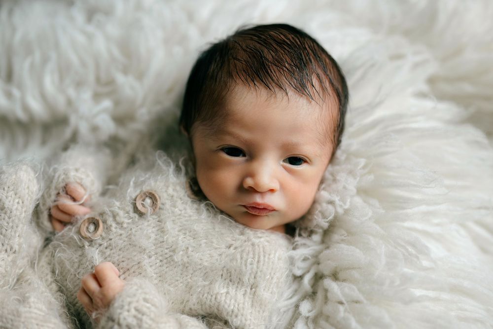 Newborn baby with dark hair lying on a fluffy white fur blanket wrapped in a soft knit cream sweater.