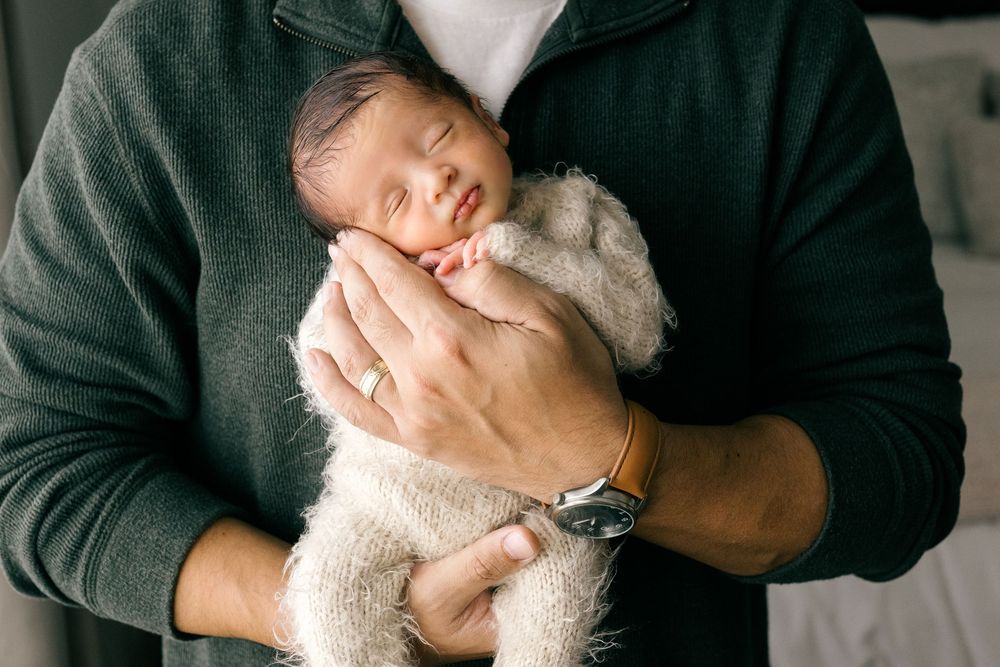 A father cradles his sleeping newborn baby in both hands, wearing a watch and dark green sweater.