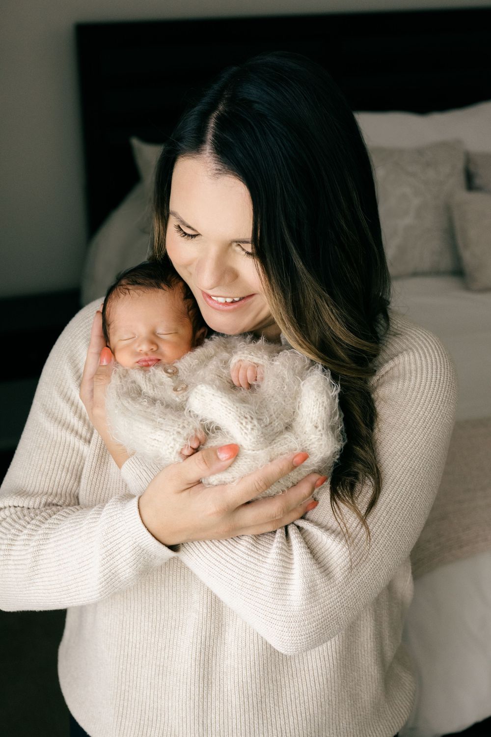 Smiling mother in cream sweater gently cradling sleeping newborn wrapped in white fuzzy blanket indoors.