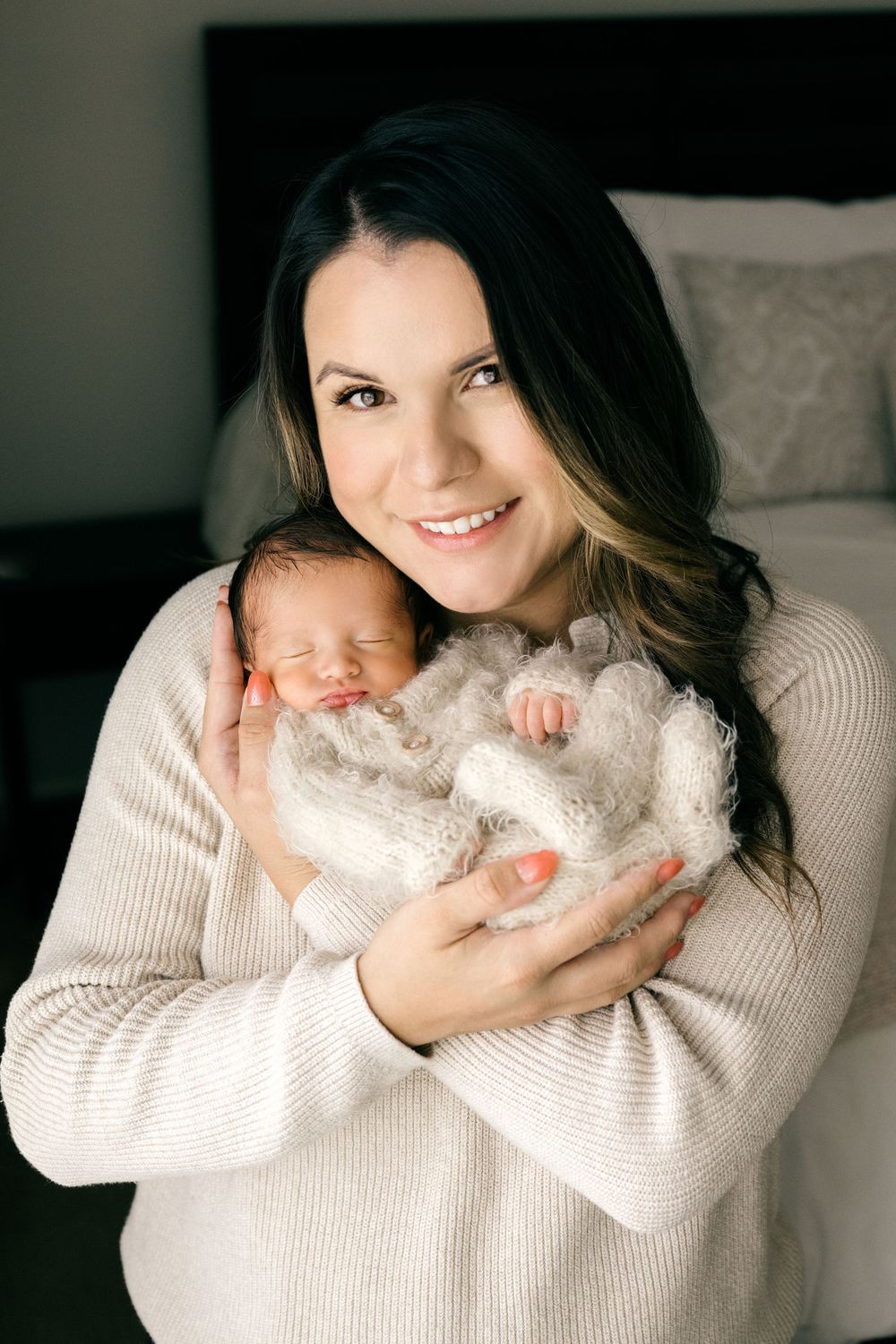 Smiling mother holding her sleeping newborn baby wrapped in a cozy white knit blanket.