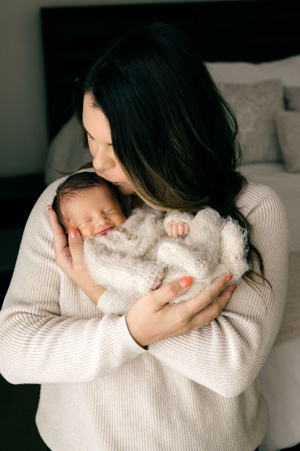 Dark-haired mother in beige sweater kissing sleeping newborn baby wrapped in soft white knit blanket.