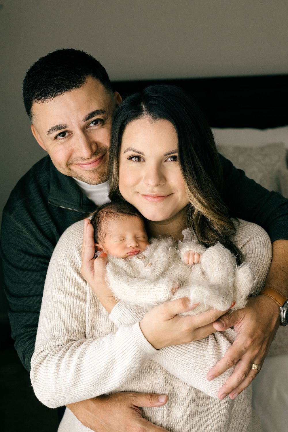 Happy parents smiling together while holding their sleeping newborn baby in a white wrap.