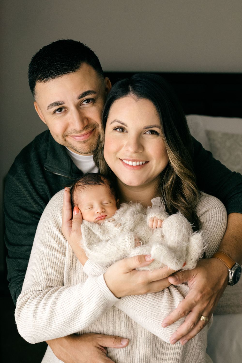 Smiling couple holding their sleeping newborn baby wrapped in white, posing together in a cozy bedroom.