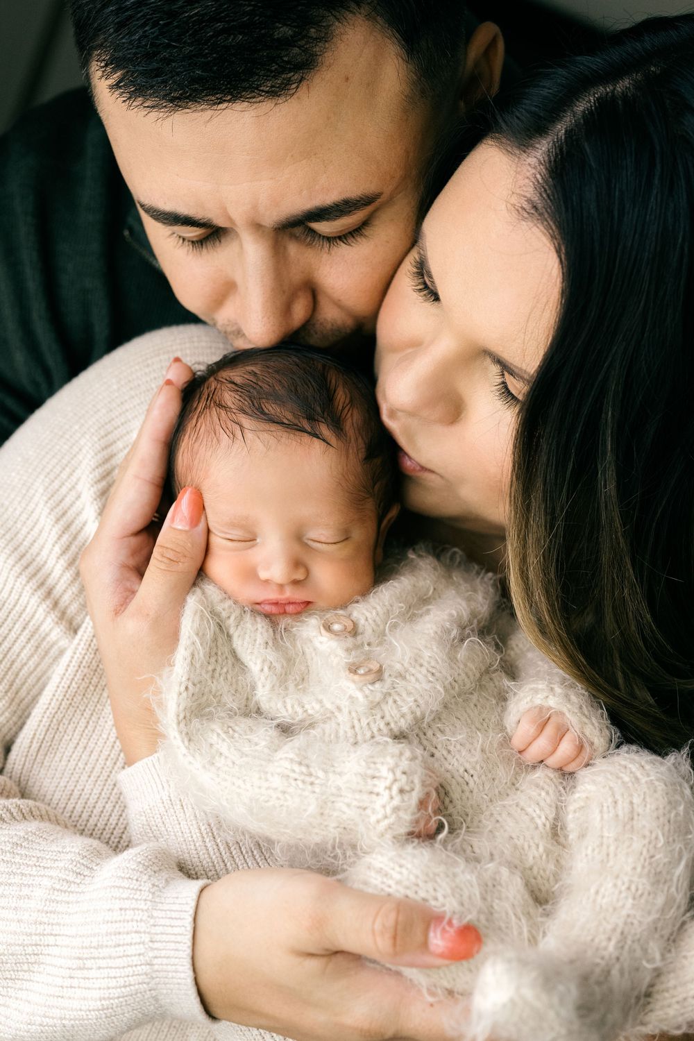 A couple tenderly kisses their sleeping newborn baby wrapped in a white knit outfit.