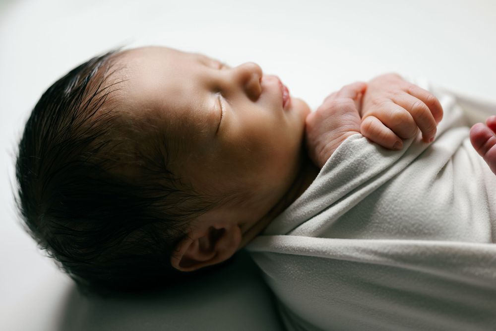 Sleeping newborn baby swaddled in white wrap with dark hair, peaceful close-up portrait.