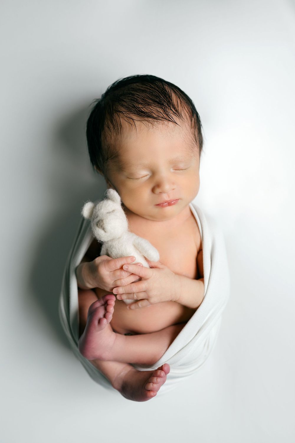 Sleeping newborn baby swaddled in white wrap holding a tiny teddy bear toy.