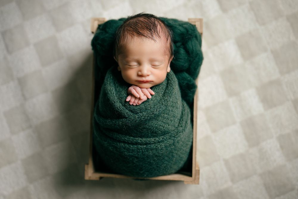 Newborn baby wrapped in green knit blanket sleeping in wooden crate, studio portrait.