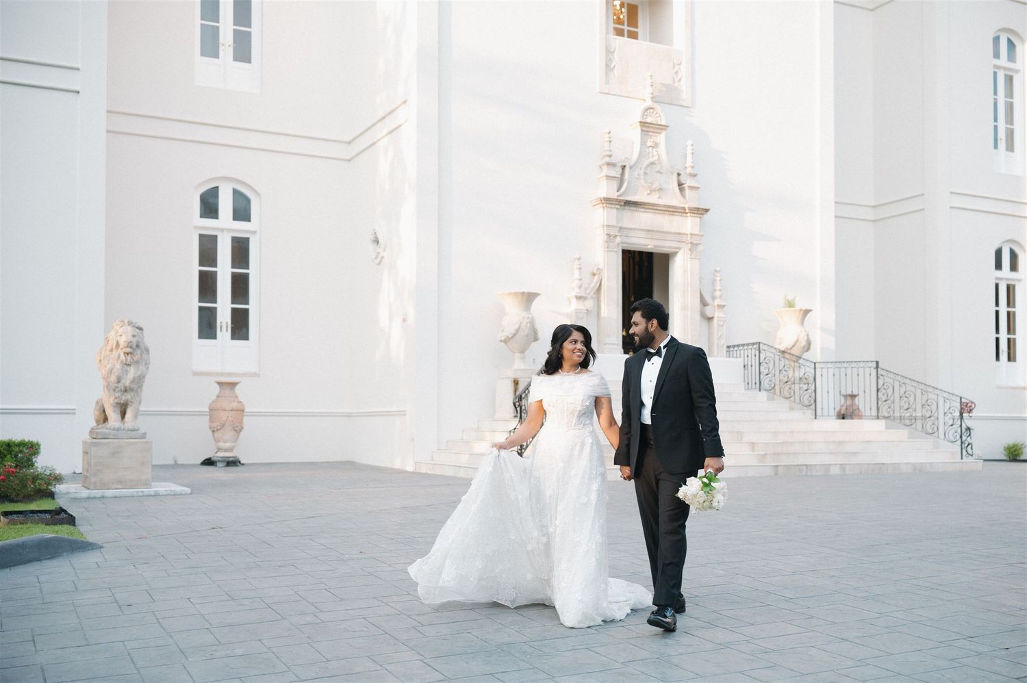 A couple walks together outside an elegant white building on their wedding day.