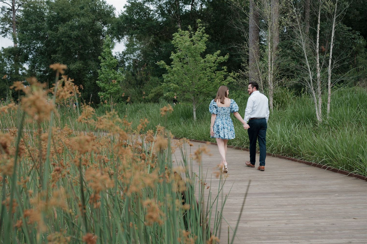 A couple walks along a wooden boardwalk path surrounded by tall grass and orange wildflowers in a natural setting.