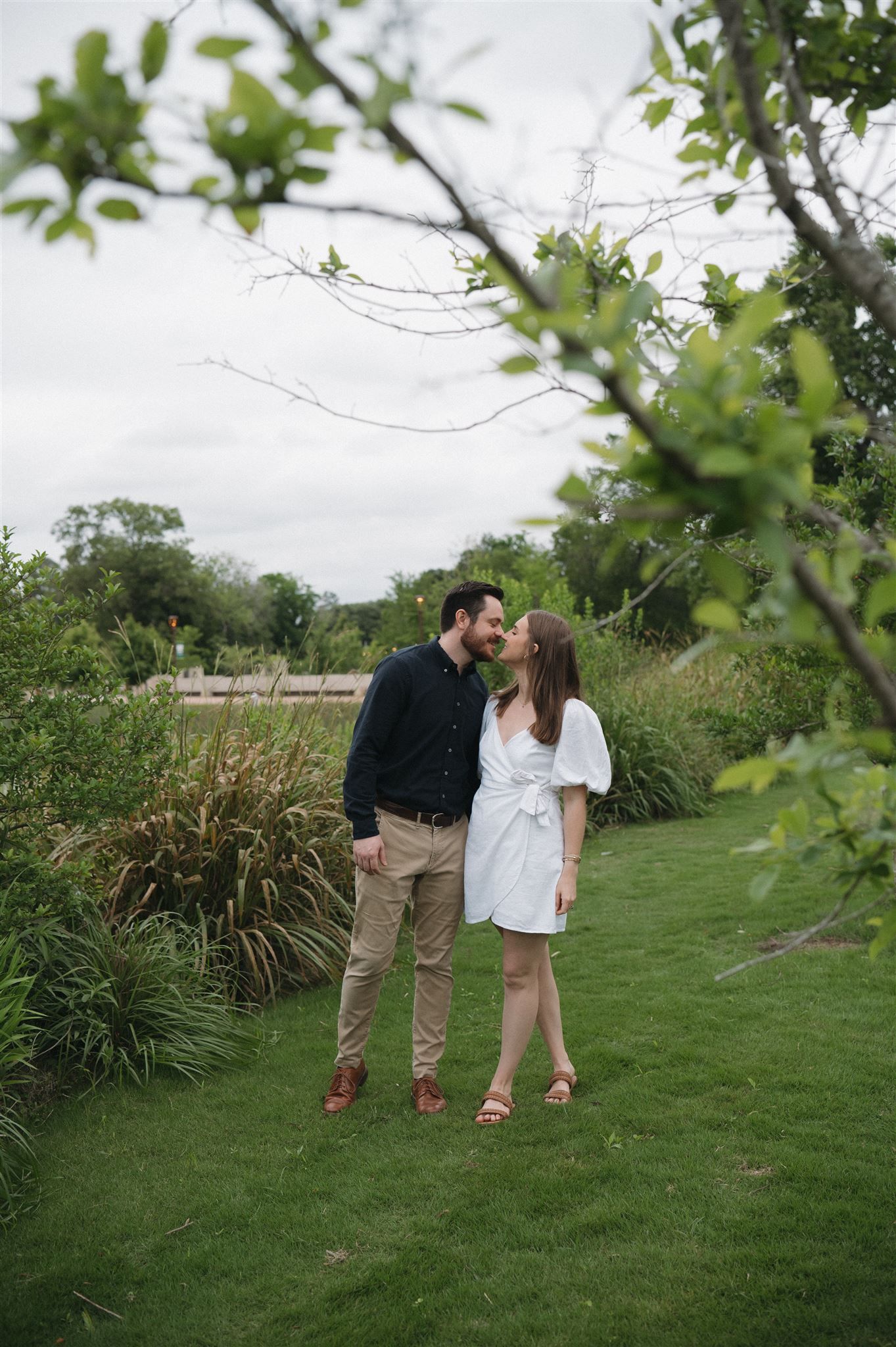 A couple shares a romantic moment on a lush green lawn surrounded by ornamental grasses and trees.