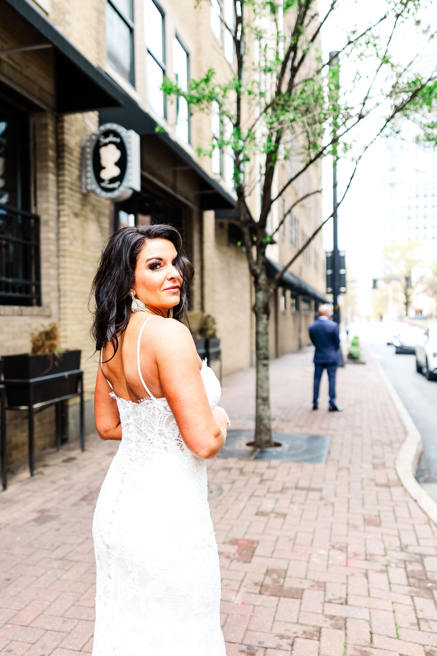 A woman in an elegant white lace wedding dress poses on a brick sidewalk in an urban setting.