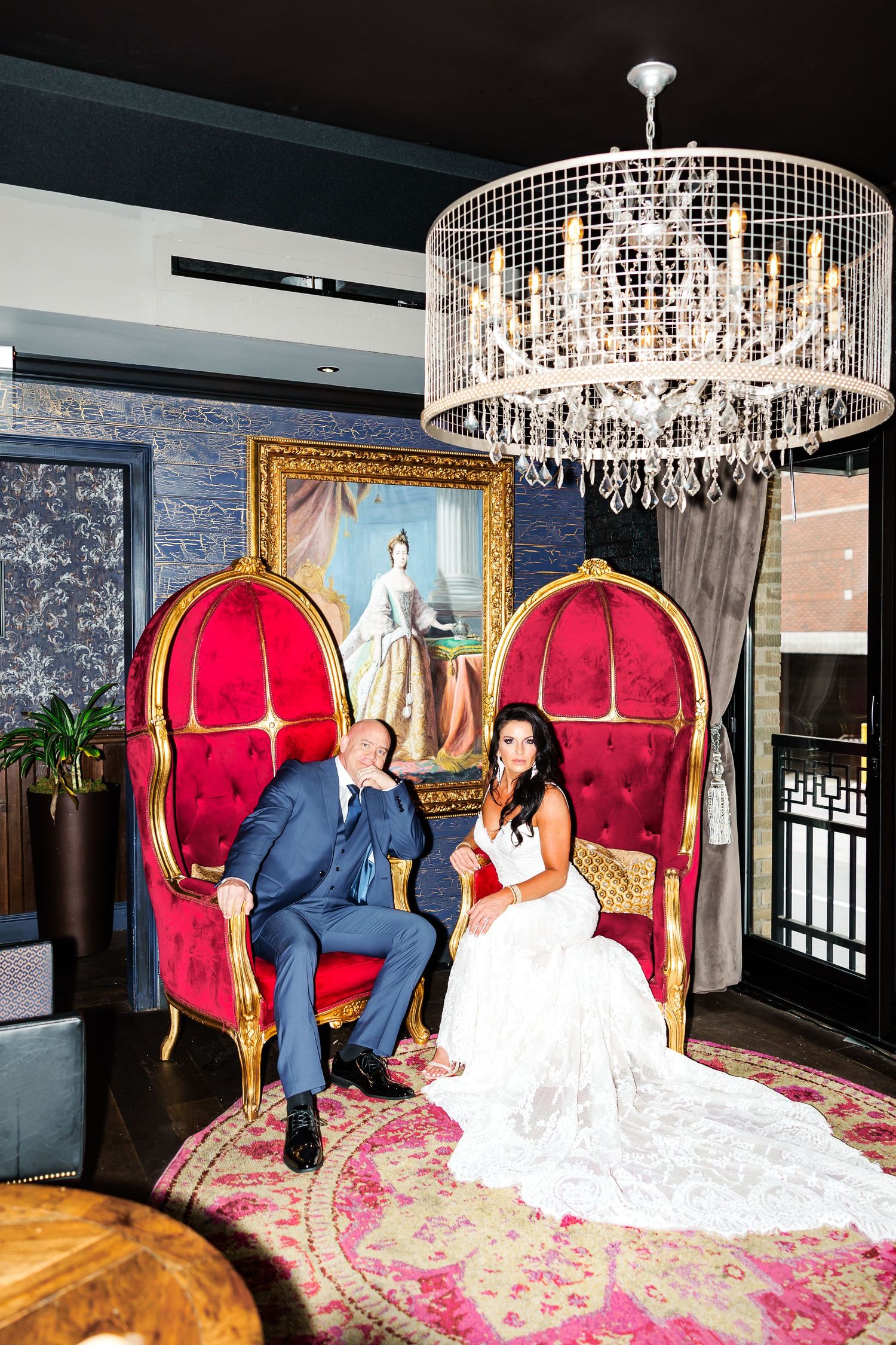 Elegant wedding couple sits on plush red velvet thrones under sparkling crystal chandeliers in a luxurious venue.