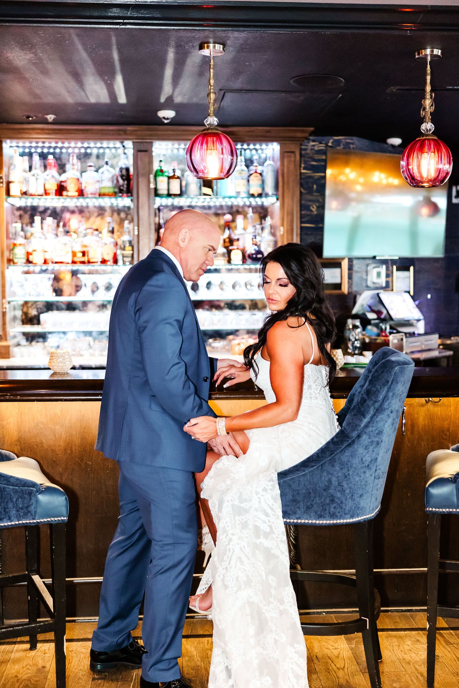 A couple shares an intimate moment at an elegant bar with red pendant lights and a fully stocked liquor display.