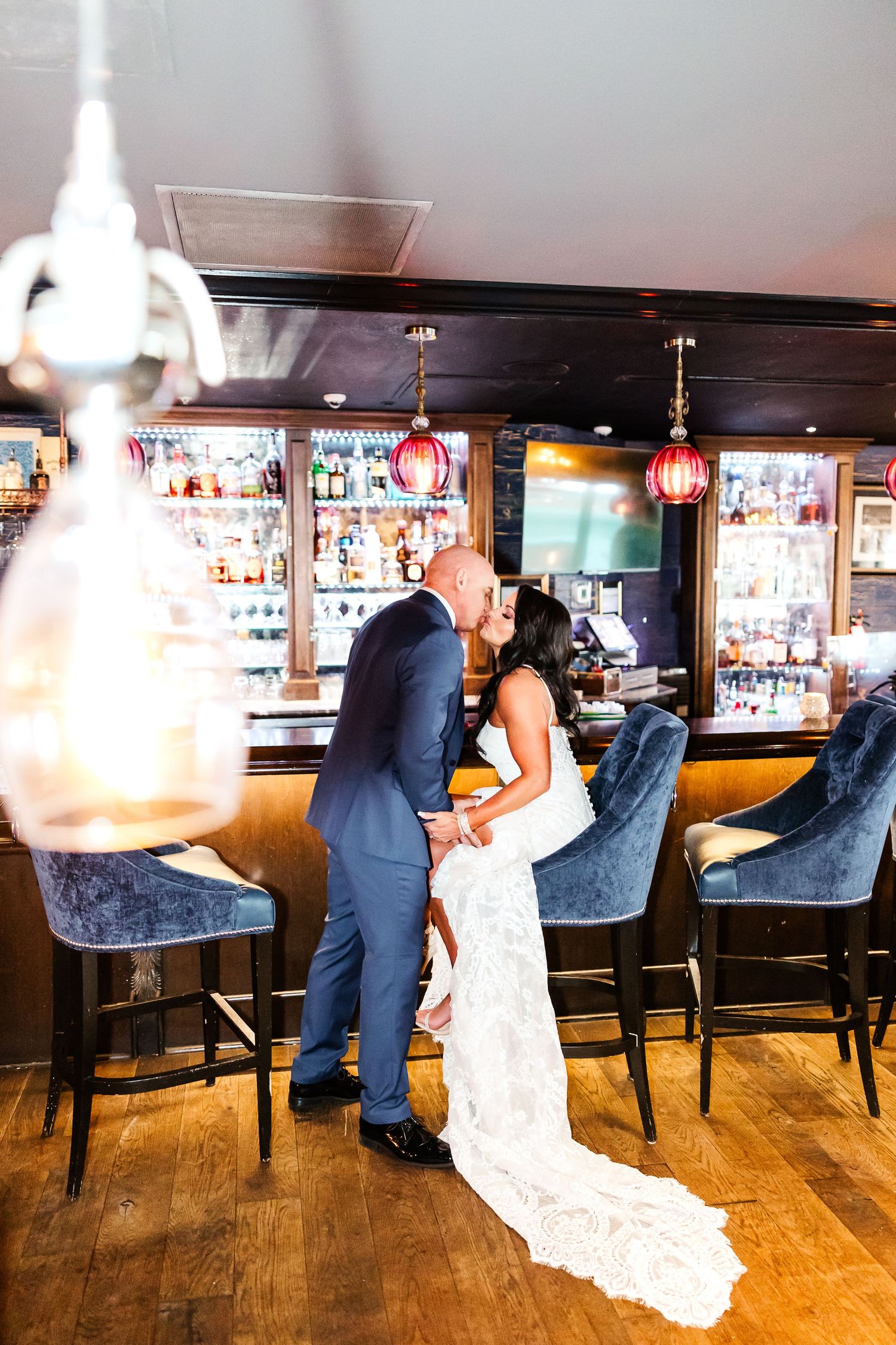 A couple in wedding attire share an intimate moment at an elegant bar with vintage pendant lighting and plush velvet seating.