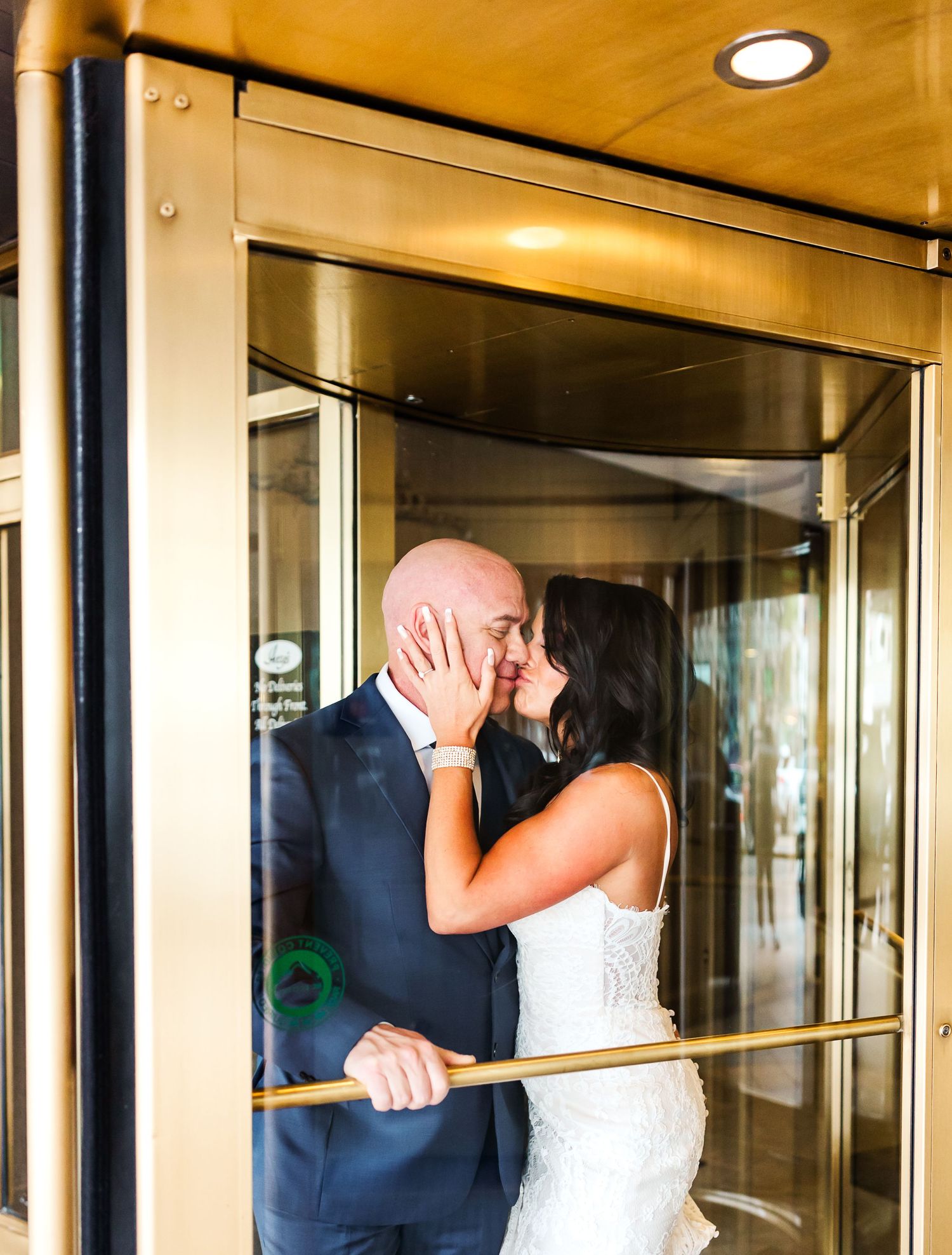 A romantic couple shares a kiss in a revolving door entrance, with the bride in white and groom in navy suit.