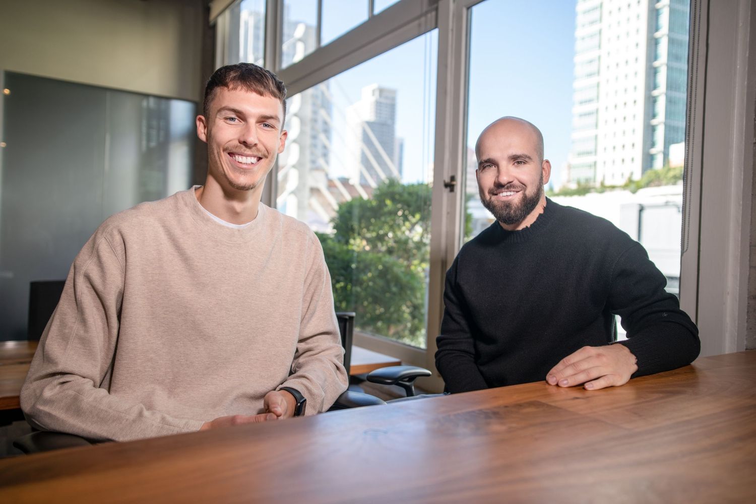 Two businessmen sitting at wooden table in bright modern office with city views through large windows.