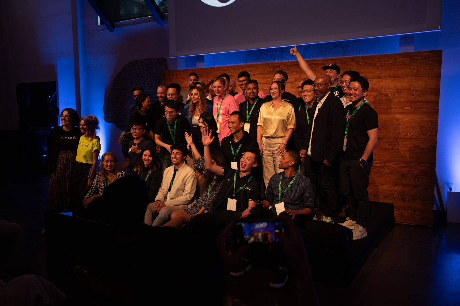A large group poses together on a stage with blue lighting and wooden backdrop during an event.