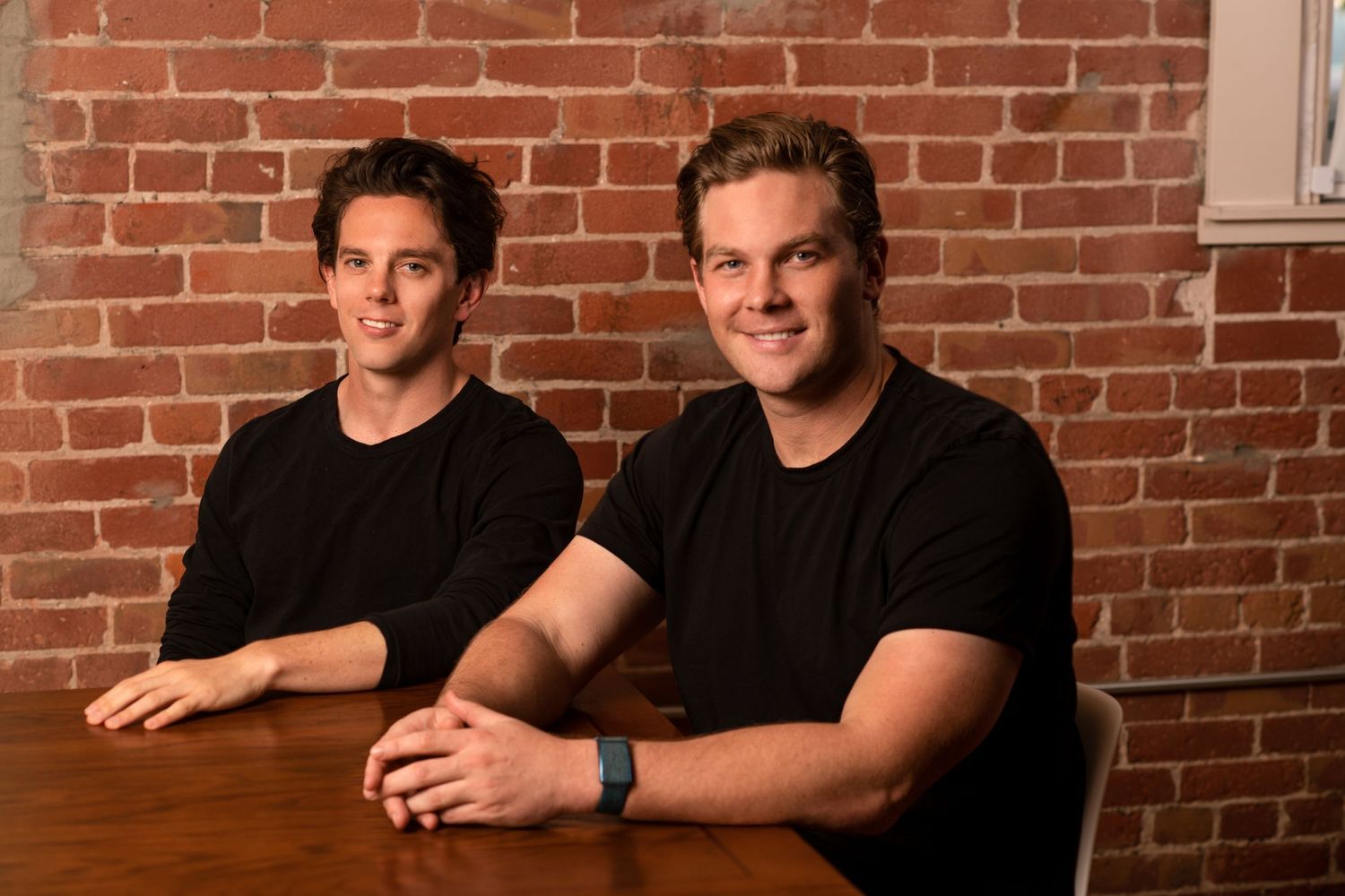 Two people wearing black t-shirts sit at a wooden table against a brick wall in casual indoor setting.