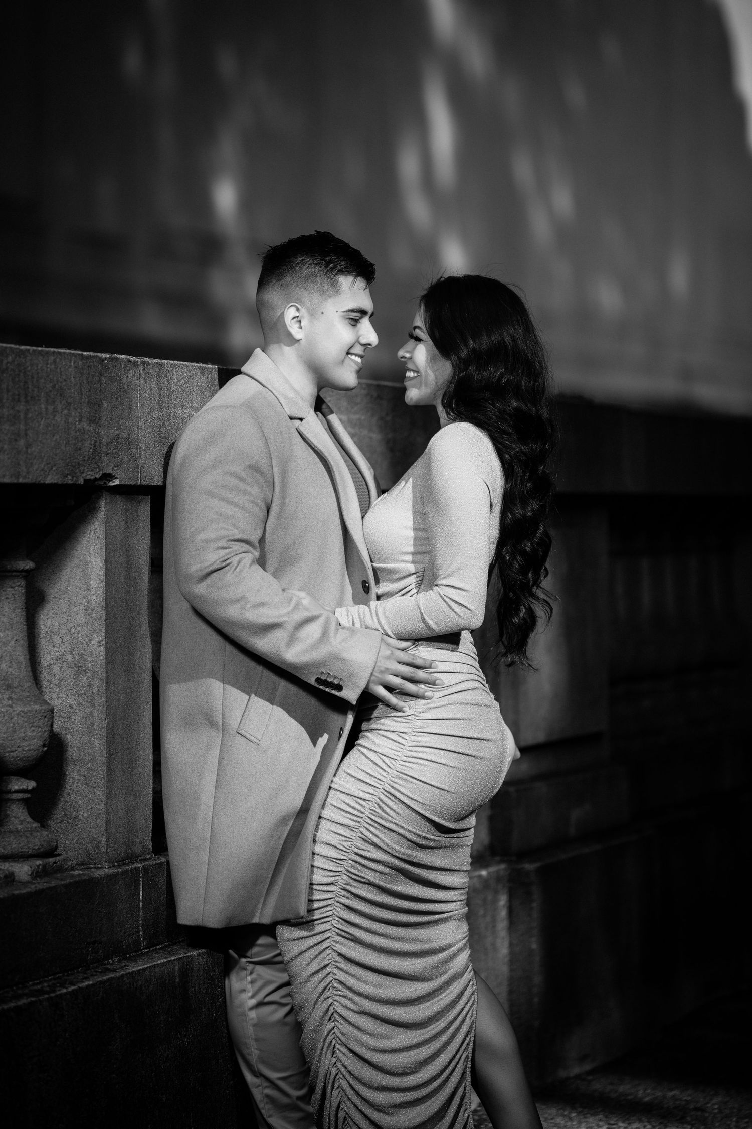 A romantic black and white portrait of a couple in formal attire embracing against a textured wall.